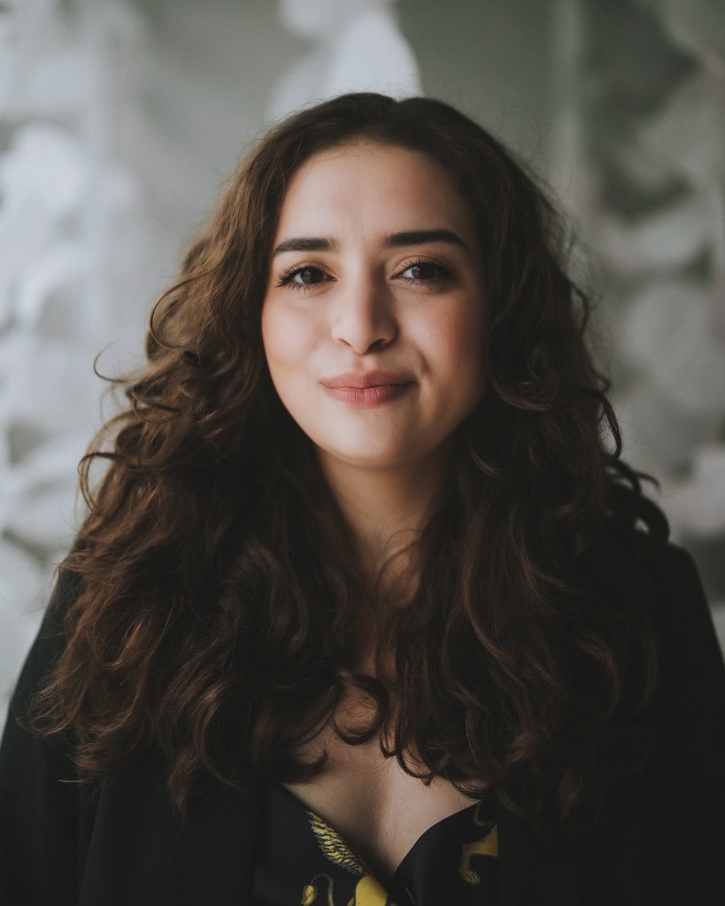A close-up portrait of a woman with curly brown hair, wearing a black blazer and a dark top. She has a warm smile and is set against a blurred background with soft lighting.