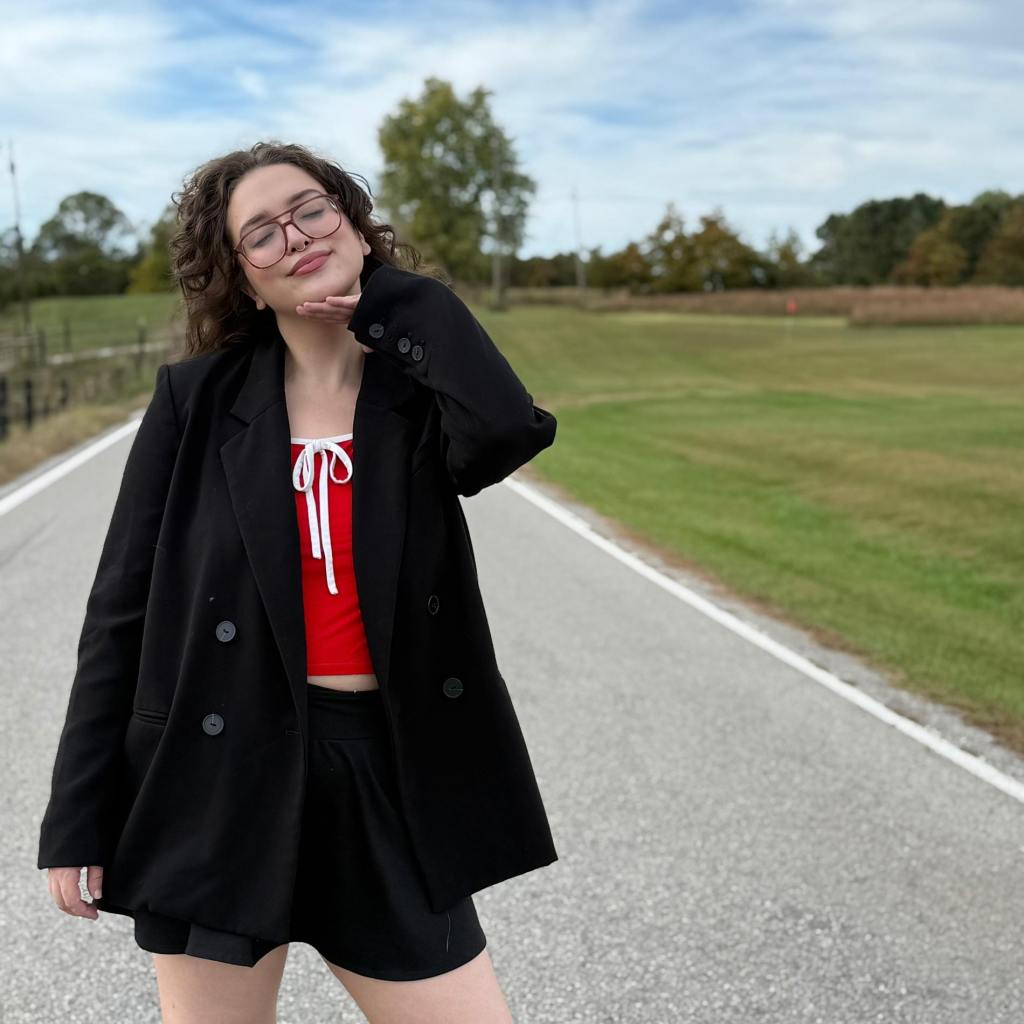 A confident individual posing playfully on a rural road, wearing a black oversized blazer over a red top and black shorts, with natural scenery in the background.